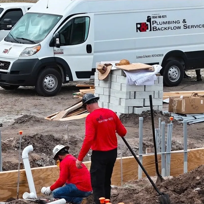 Two R&J plumbers in branded red shirts setting underground rough-in pipe at a new construction slab, service van behind them